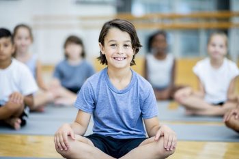 A group of elementary school children are indoors in the gym. A boy in the foreground is smiling at the camera while sitting crosslegged.